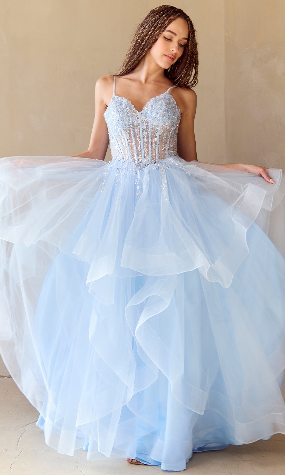 A woman stands indoors against a beige wall, wearing the Amelia Couture 5068 Long Glitter Prom Ball Gown—a light blue, layered tulle dress with a fitted, embellished bodice and spaghetti straps.